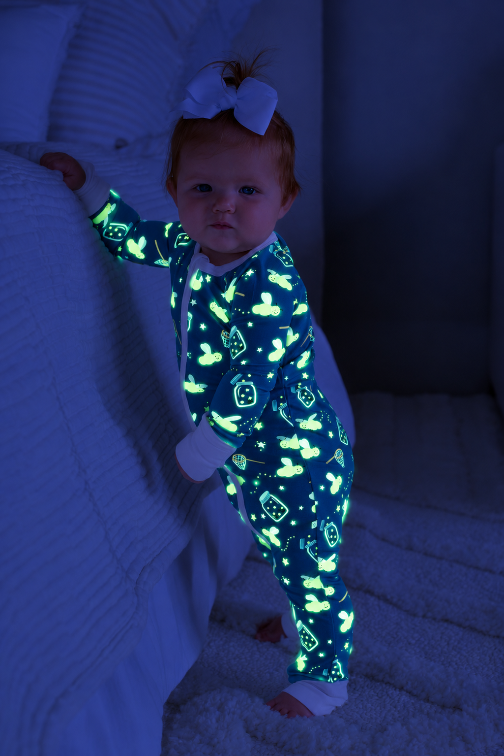 Baby standing, holding onto a bed, wearing a Caden Lane blue convertible bamboo zip romper in a Chasing Fireflies print featuring fireflies and mason jars, paired with a white bow.