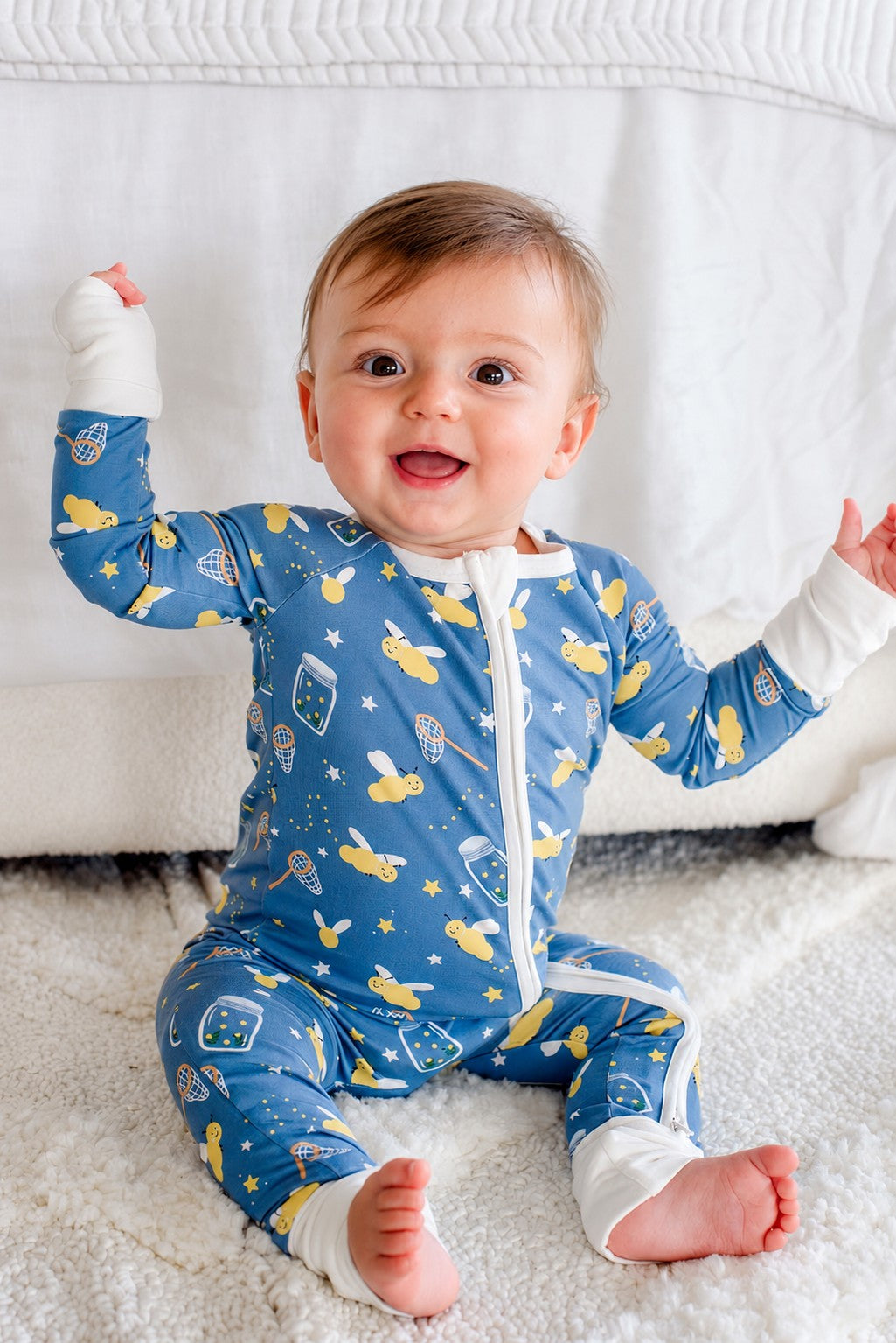 Side view of a baby boy sitting on a bed wearing a Caden Lane blue convertible bamboo zip romper in a Chasing Fireflies print featuring fireflies and mason jars.