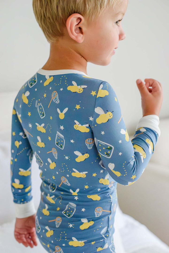 Back view of a toddler boy wearing a Caden Lane blue two-piece bamboo pajama set in a Chasing Fireflies print featuring fireflies and mason jars, with cream cuffs and collar.