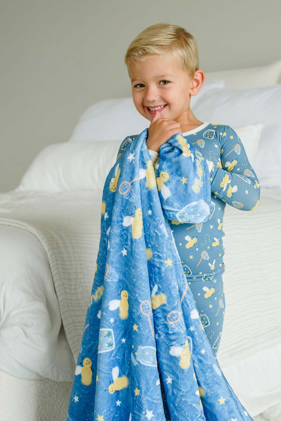 Smiling toddler boy standing on a bed holding up a Caden Lane blue plush personalized blanket in a Chasing Fireflies print featuring fireflies and mason jars, paired with matching pajamas.