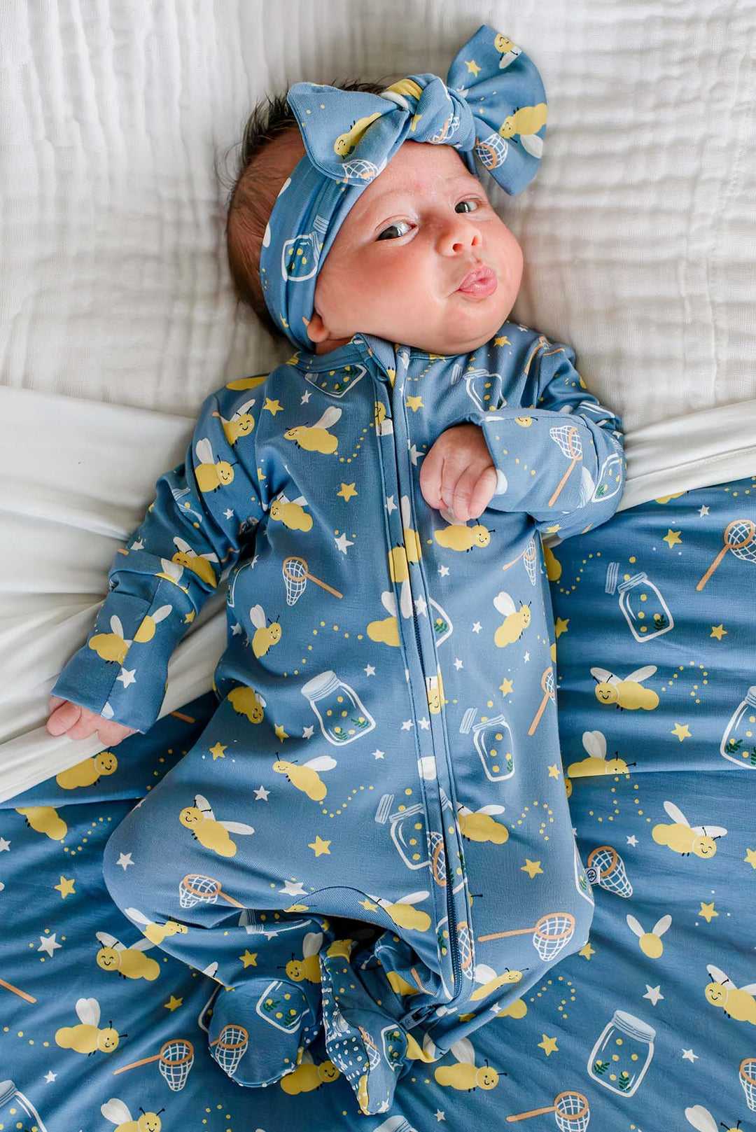 Overhead shot of a newborn wearing a Caden Lane blue knit large bow headwrap in a Chasing Fireflies print featuring fireflies and mason jars, paired with a matching zipper footie.