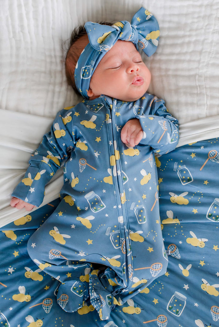 Newborn stretching her arms, wearing a Caden Lane blue knit large bow headwrap in a Chasing Fireflies print featuring fireflies and mason jars, paired with a matching zipper footie.