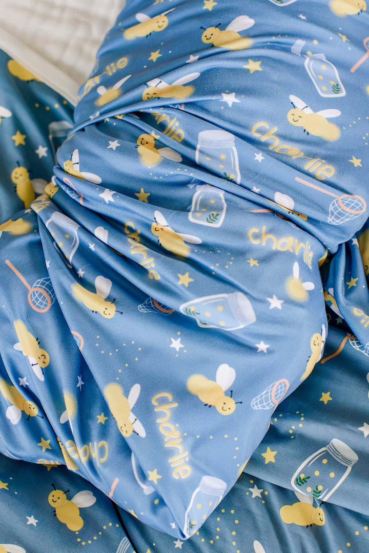 Close-up of the folded layers of a Caden Lane blue personalized swaddle in a Chasing Fireflies print featuring fireflies and mason jars, reading "Charlie" in yellow script.