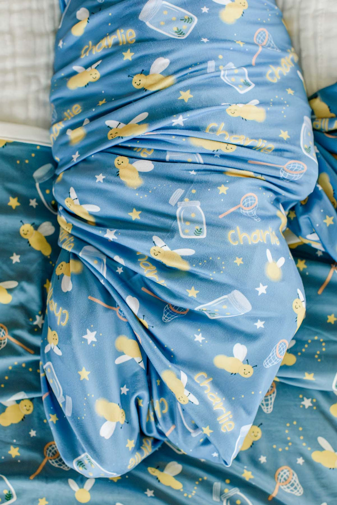 Close-up of a baby wrapped snugly in a Caden Lane blue personalized swaddle in a Chasing Fireflies print featuring fireflies and mason jars, reading "Charlie."