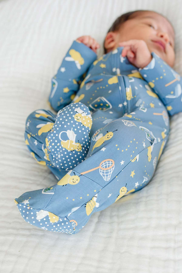 Newborn lying with knees raised to show the polka-dot soles of a Caden Lane blue bamboo zipper footie in a Chasing Fireflies print featuring fireflies and mason jars.