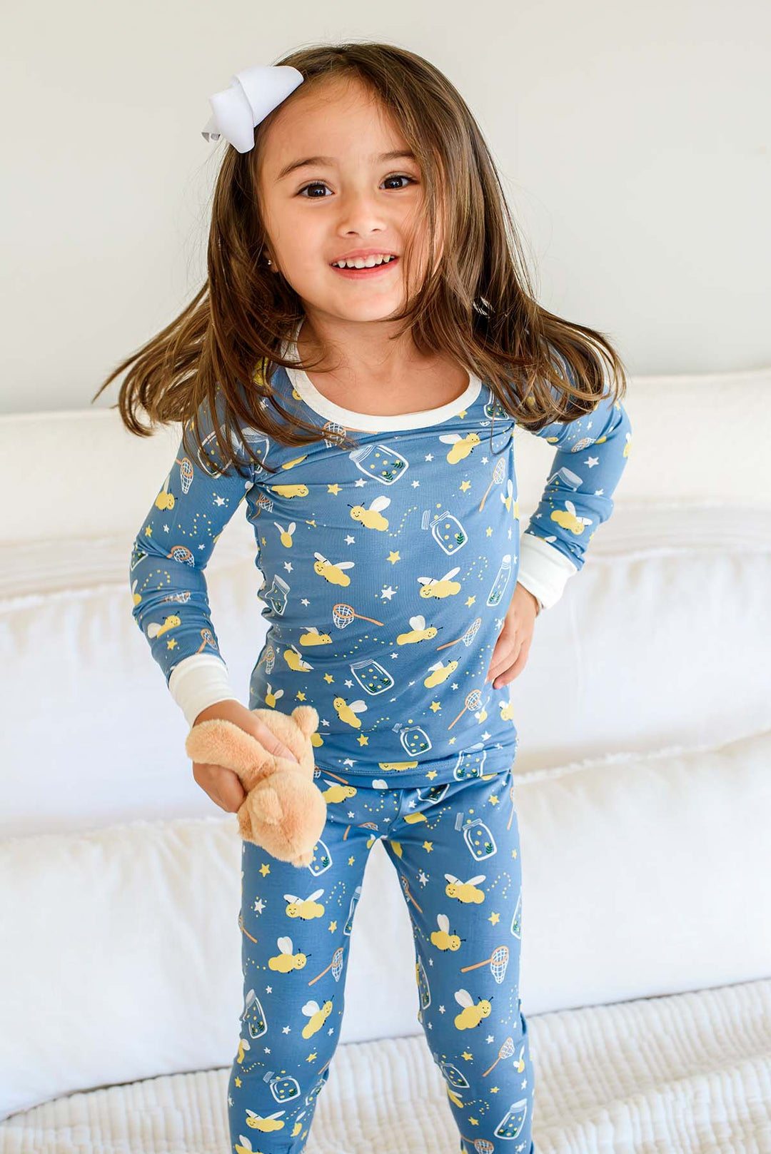 Girl posing, hand on hip, wearing a Caden Lane blue two-piece bamboo pajama set in a Chasing Fireflies print featuring fireflies and mason jars, paired with a teddy bear.