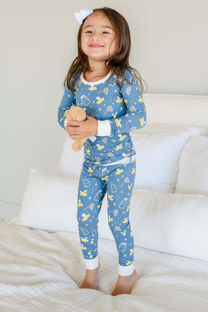 Full-length shot of a smiling girl wearing a Caden Lane blue two-piece bamboo pajama set in a Chasing Fireflies print featuring fireflies and mason jars, paired with a teddy bear.