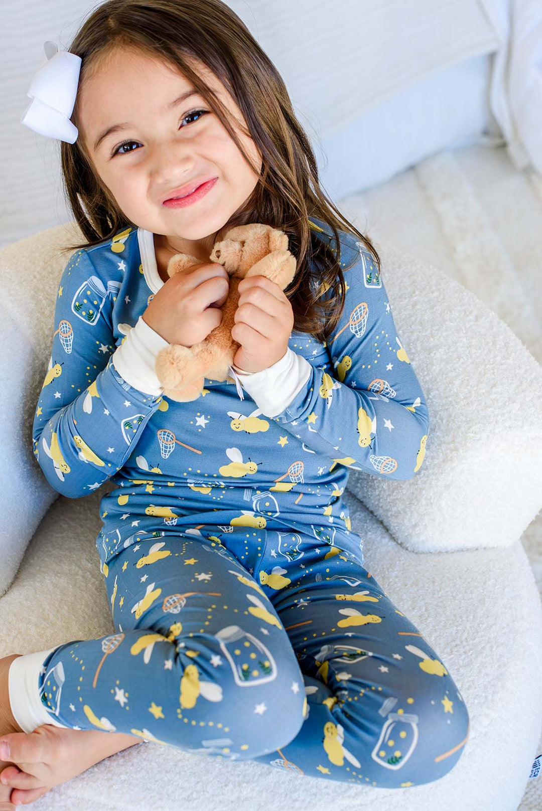 Grinning girl hugging a teddy bear, wearing a Caden Lane blue two-piece bamboo pajama set in a Chasing Fireflies print featuring fireflies and mason jars, paired with a white bow.