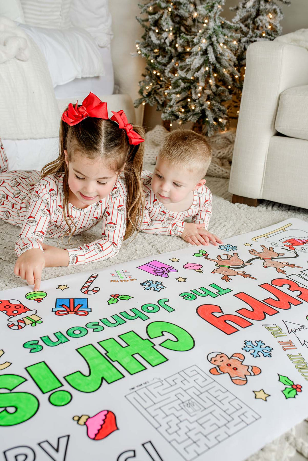 Girl with red bow and toddler brother in Santa pajamas pointing at a personalized Merry Christmas coloring banner by the tree