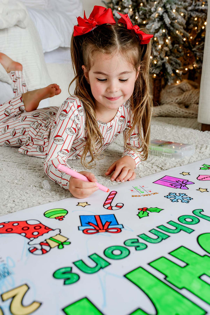 Smiling girl in Santa pajamas coloring a candy cane with a pink marker on a personalized Christmas coloring banner
