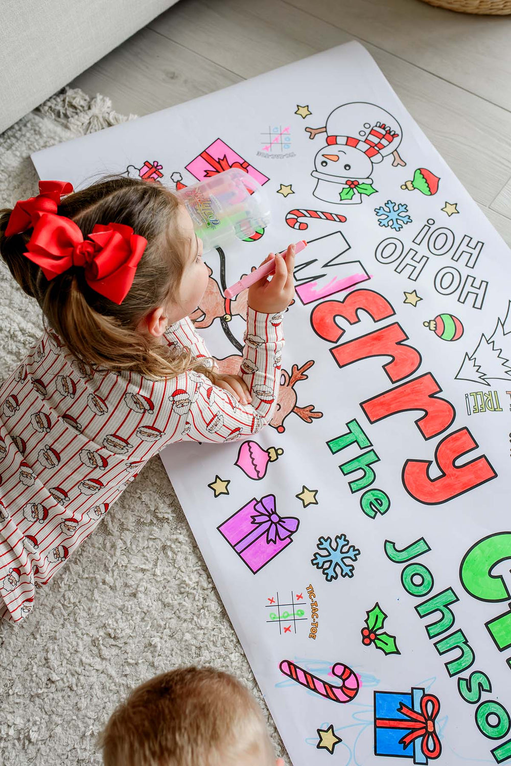 Girl with red bow coloring the word Merry in red on a personalized Christmas coloring banner with snowman and candy canes