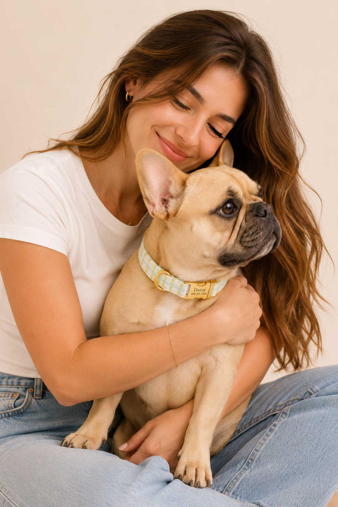 Tan French Bulldog seated and looking up, wearing a picnic plaid dog collar with gold "Daisy" ID tag