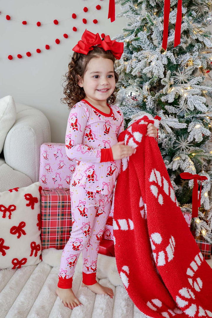 Toddler girl in pink Santa and bow print pajamas holding a Caden Lane red candy cane print sherpa blanket in front of a Christmas tree