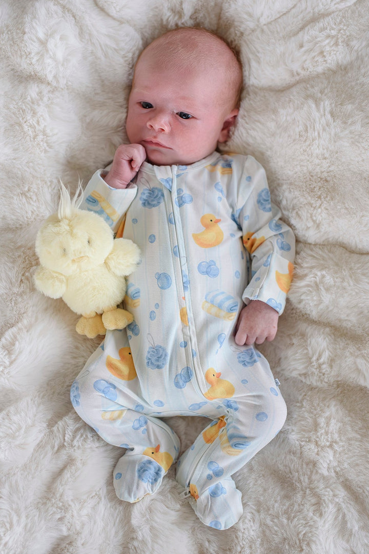 Newborn lying on white fur wearing blue Bubble Duckies bamboo zipper footie pajamas with rubber duck stuffed toy