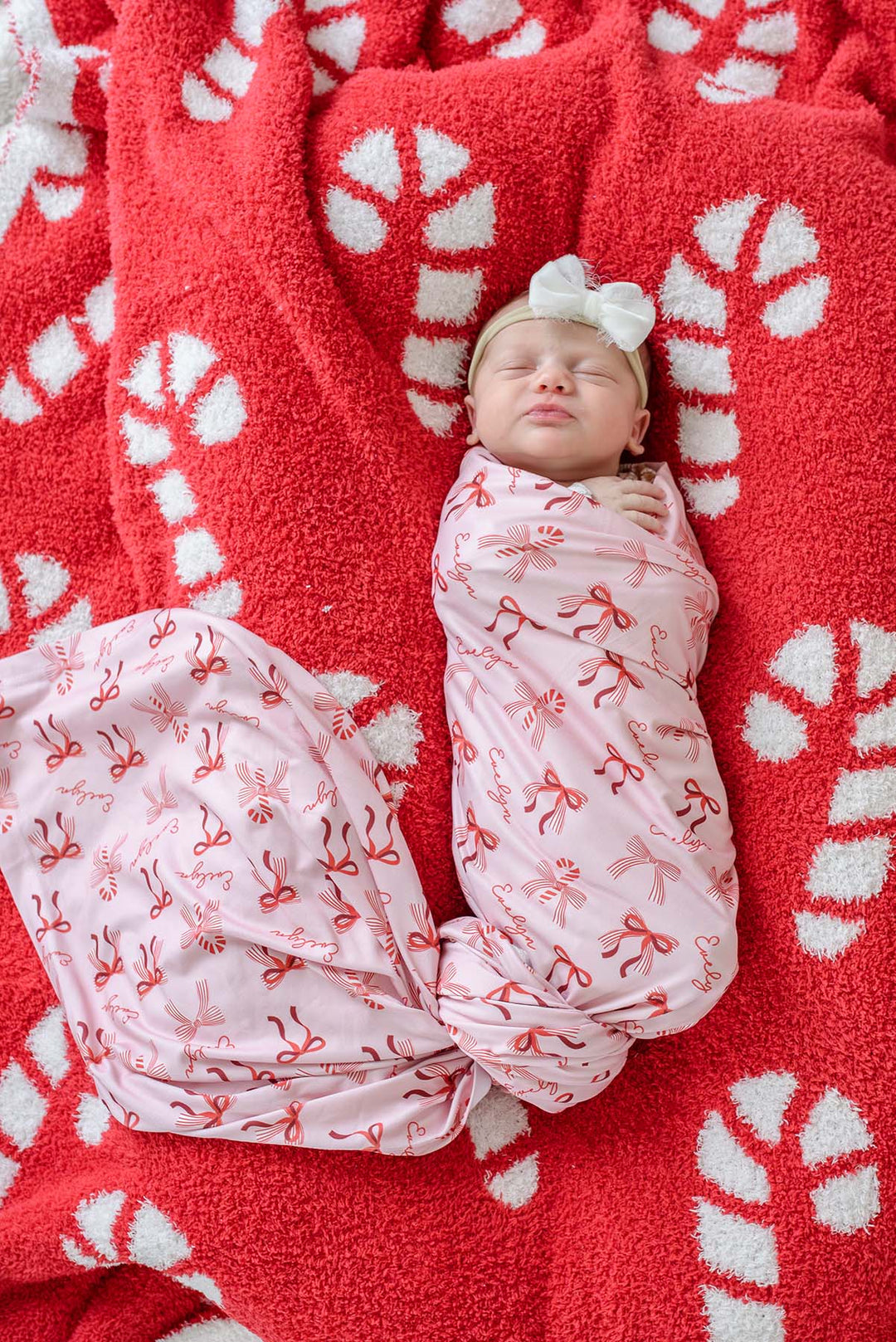 Caden Lane personalized pink bow print swaddle blanket wrapped around a sleeping newborn, laid on a red and white candy cane print sherpa blanket