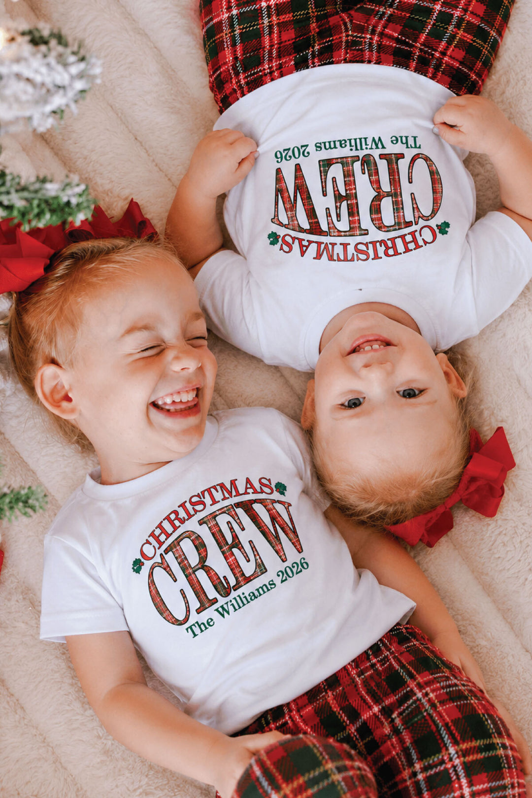 Two kids wearing personalized Caden Lane Christmas Crew shirts with coordinating red plaid pajama pants and festive red hair bows.