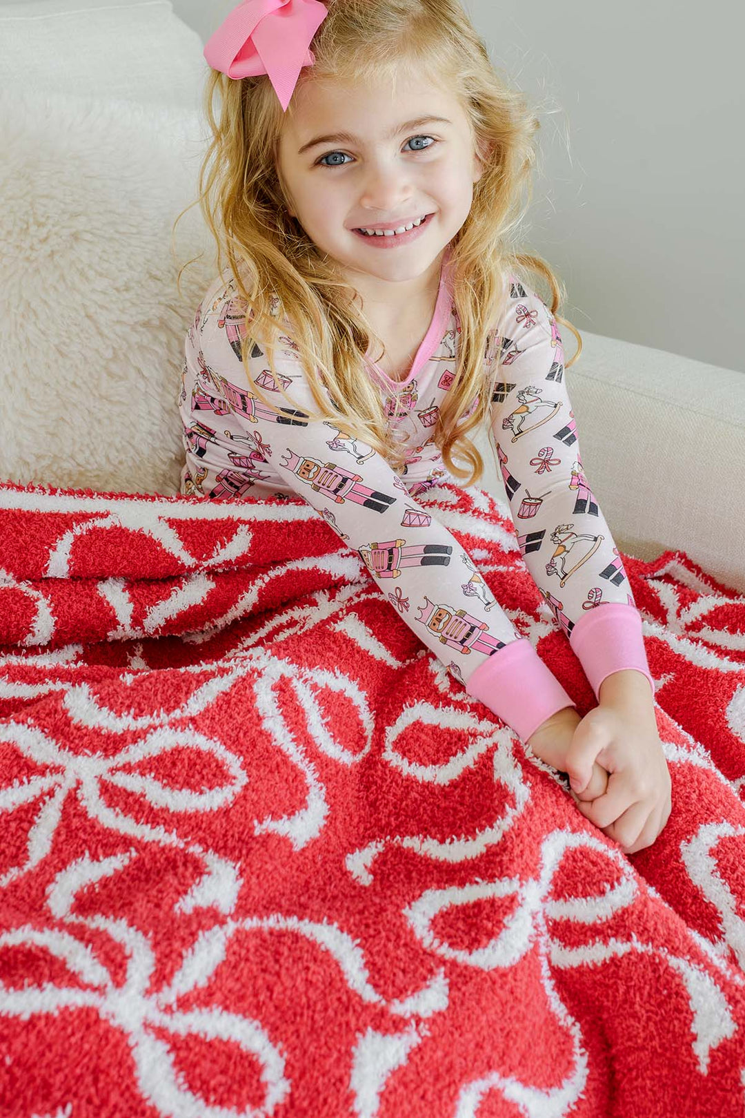 Caden Lane red bow print sherpa blanket laid over a young girl wearing pink nutcracker-print pajamas, showcasing the plush texture and allover ribbon bow pattern.
