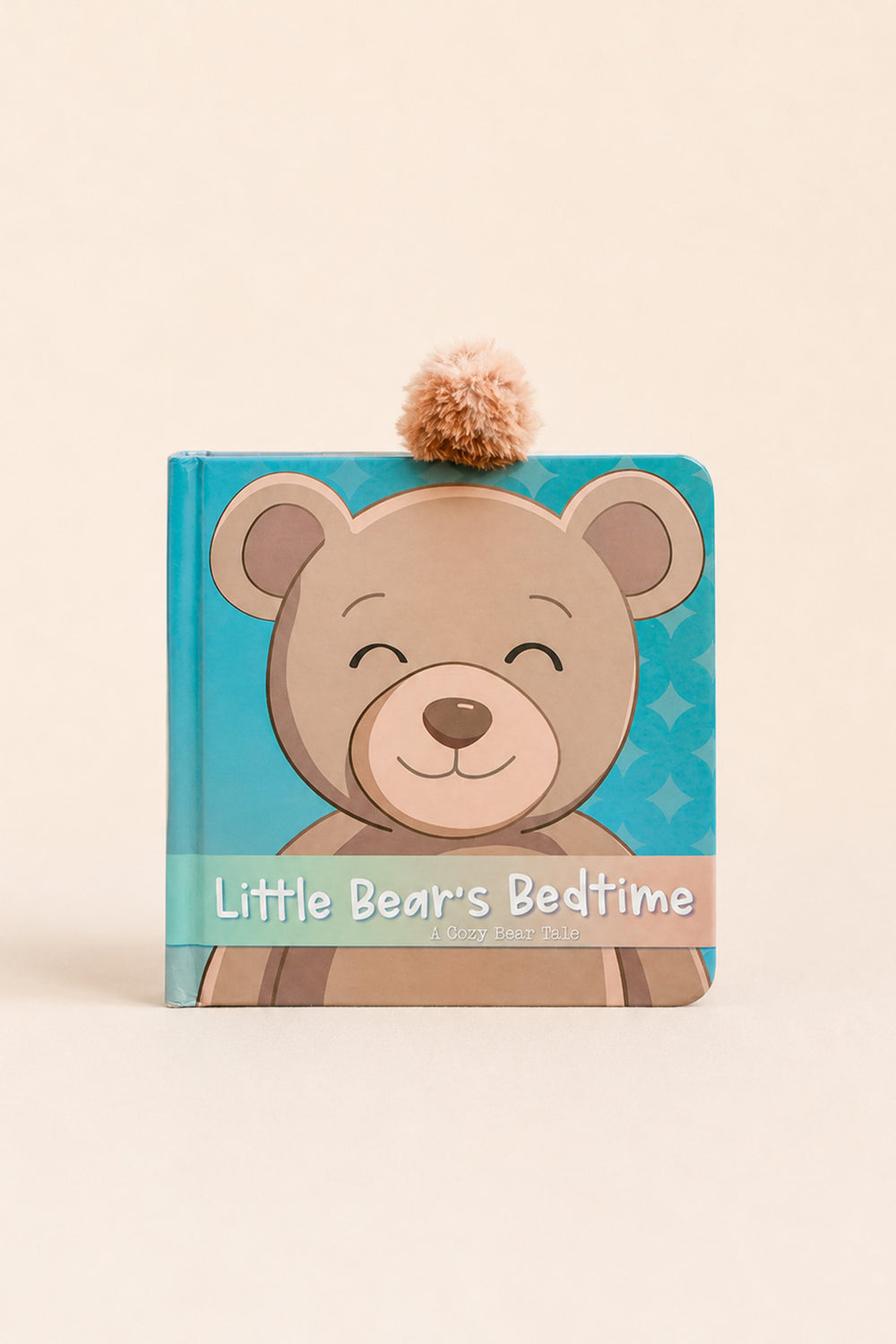 A children's hardcover book titled 'Little Bear's Bedtime' featuring a cartoon teddy bear on a turquoise cover with a small tan pom-pom on top, photographed against a warm cream background.