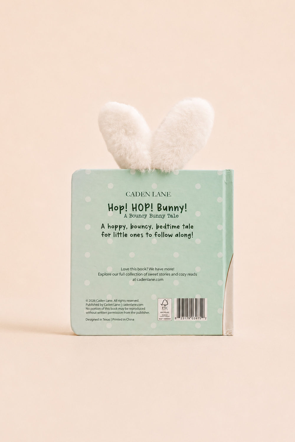 The back cover of the children's book 'Hop! Hop! Bunny!' by Cadence Lane, featuring a mint green background with white polka dots and a fluffy white bunny ear bookmark attached at the top, set against a solid pale peach background.