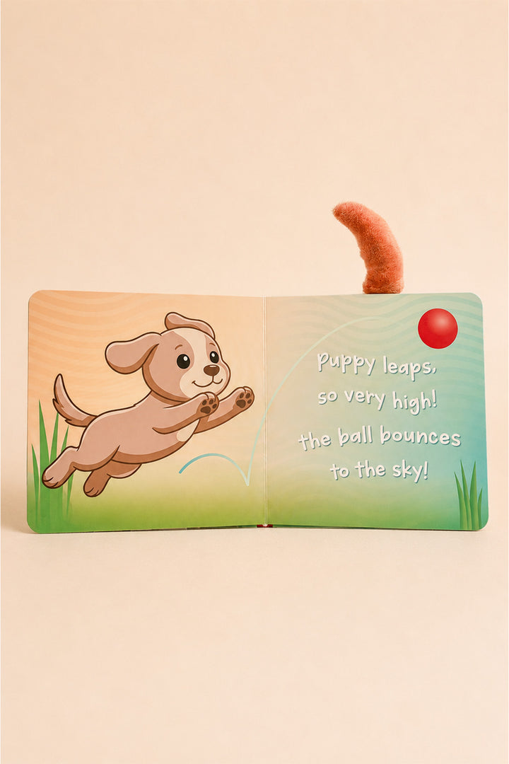 A children's pop-up book shown open on a warm beige background. The left page features an illustration of a puppy jumping, while the right page has text reading 'Puppy leaps, so very high! the ball bounces to the sky!' and includes a small pop-up tail and red ball.