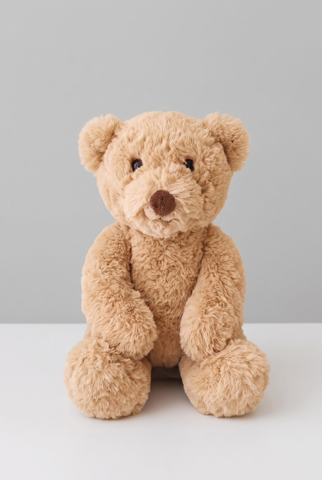 A classic plush teddy bear in a warm tan color, sitting upright against a seamless off-white background.