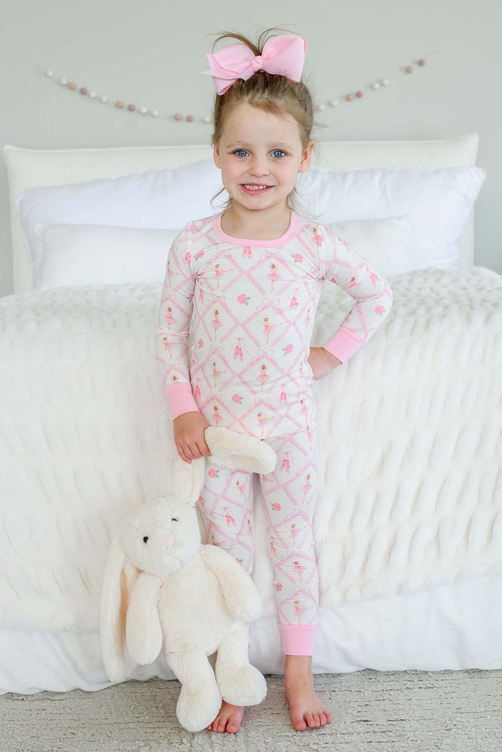 Smiling toddler girl wearing a Caden Lane ballerina and roses pink print two-piece pajama set with pink cuffs, holding a stuffed bunny while standing next to a bed