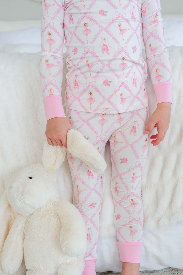 Toddler girl wearing a Caden Lane ballerina and roses pink print two-piece pajama set with pink cuffs, holding a stuffed bunny while standing next to a bed