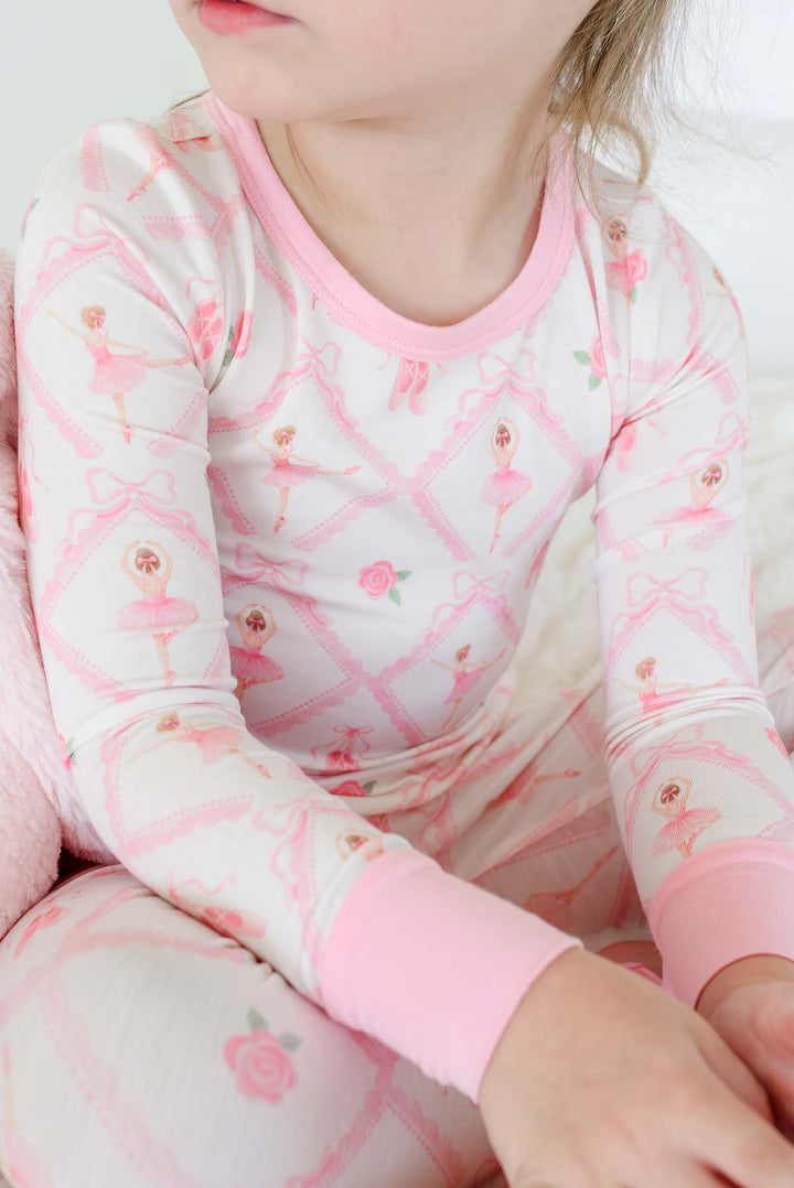 Smiling toddler girl wearing a Caden Lane ballerina and roses pink print two-piece pajama set with pink cuffs, while sitting on a bed