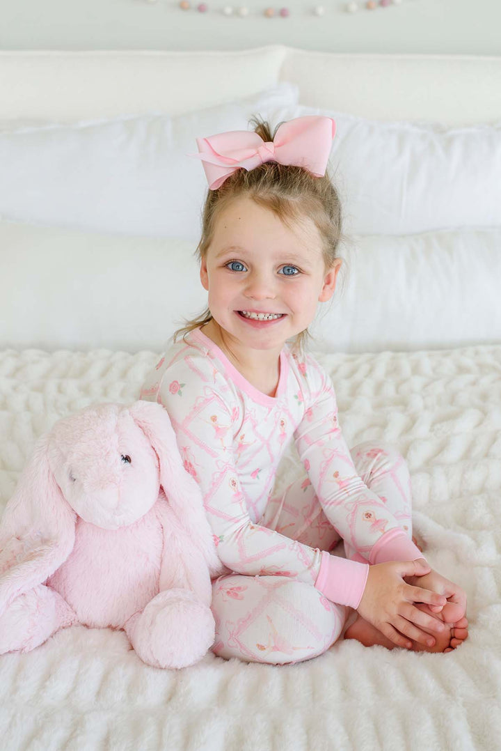 Smiling toddler girl wearing a Caden Lane ballerina and roses pink print two-piece pajama set with pink cuffs, holding a stuffed bunny while sitting on a bed