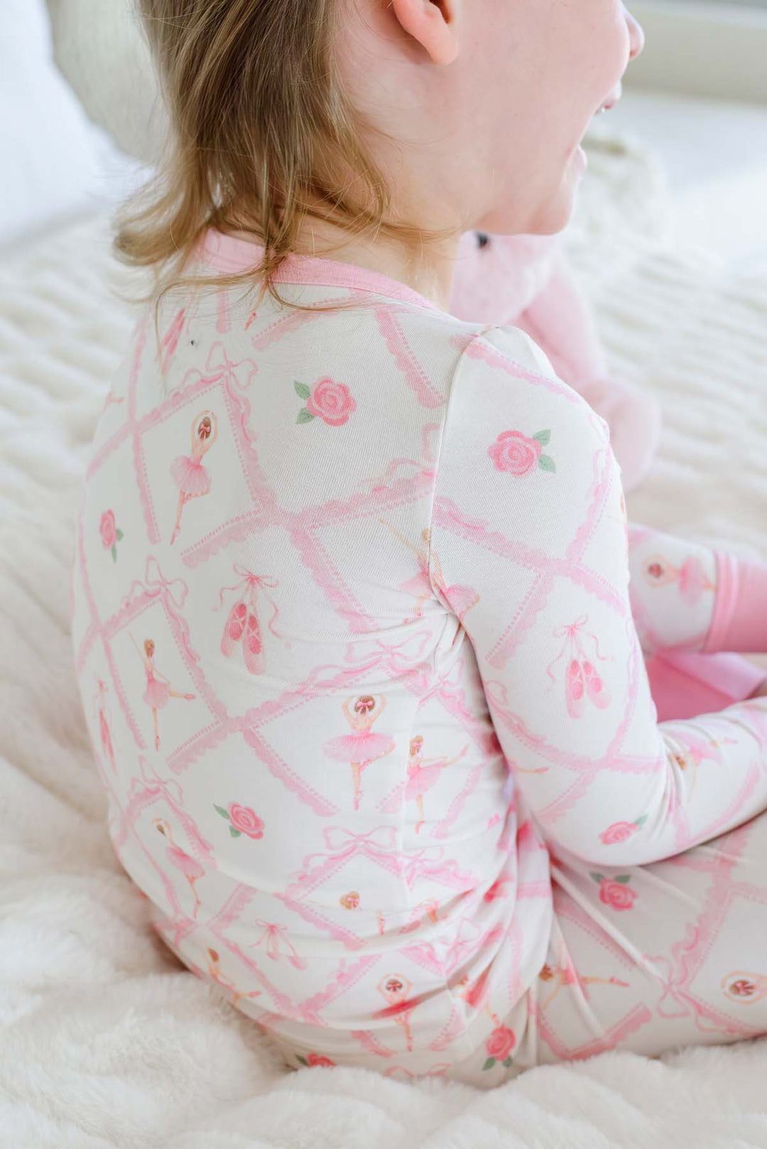 Smiling toddler girl wearing a Caden Lane ballerina and roses pink print two-piece pajama set with pink cuffs, while sitting on a bed