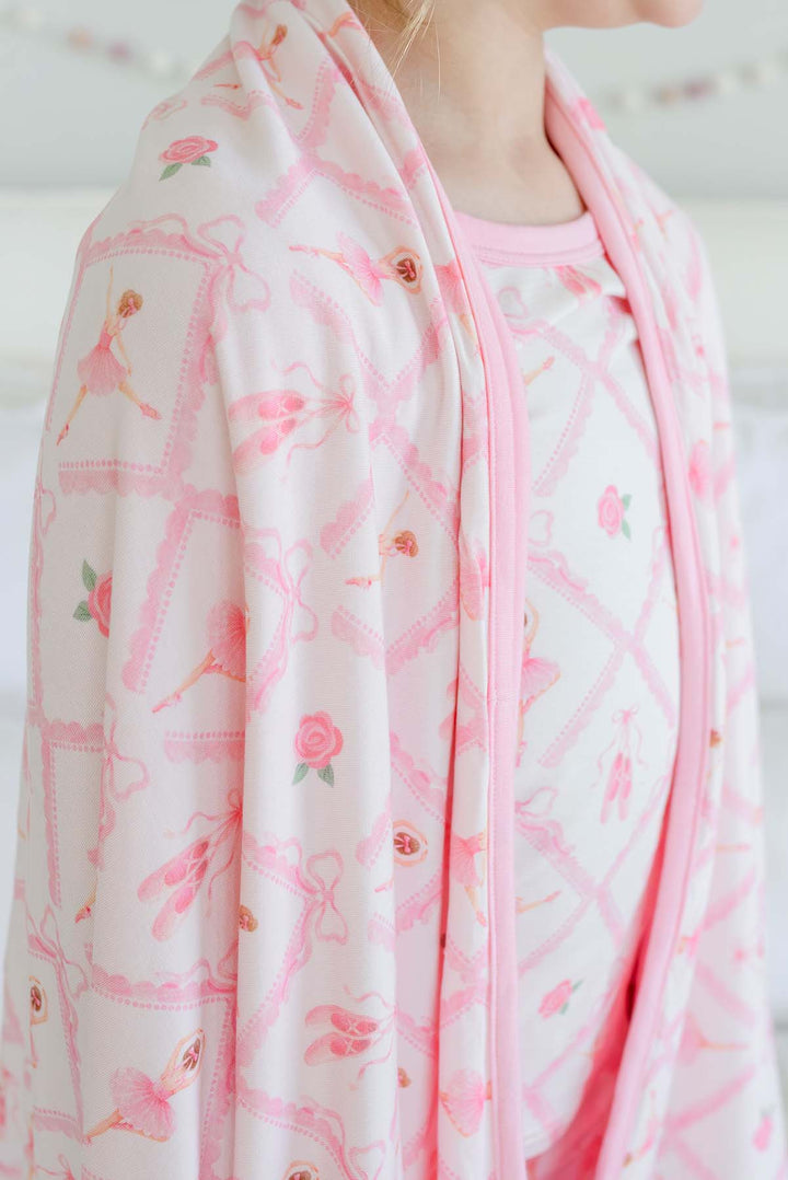 Girl standing on a bed with a pink bow in her hair, wrapped in a Caden Lane ballerina and roses pink print bamboo blanket