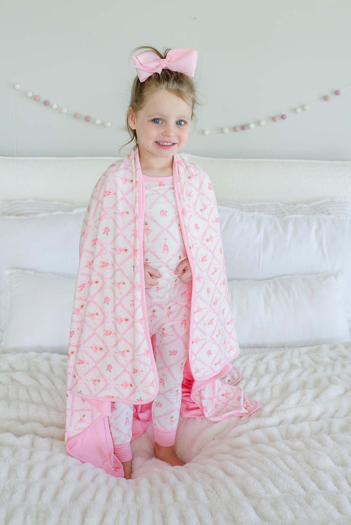 Smiling young girl standing on a bed with a pink bow in her hair, wrapped in a Caden Lane ballerina and roses pink print bamboo blanket