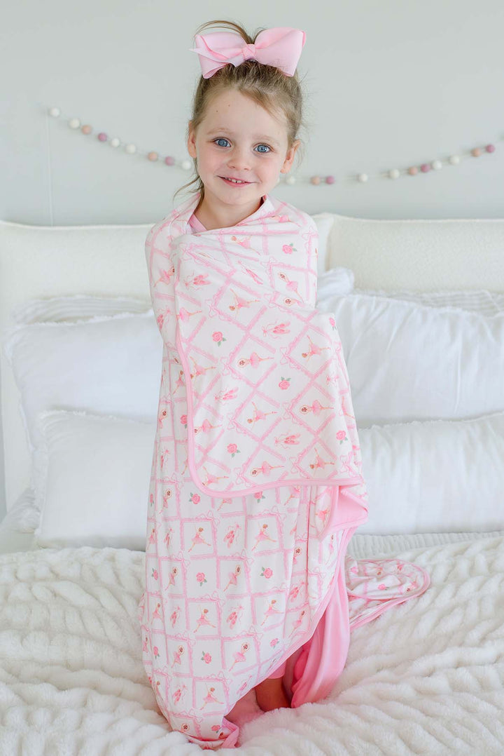 Smiling young girl standing on a bed with a pink bow in her hair, wrapped in a Caden Lane ballerina and roses pink print bamboo blanket