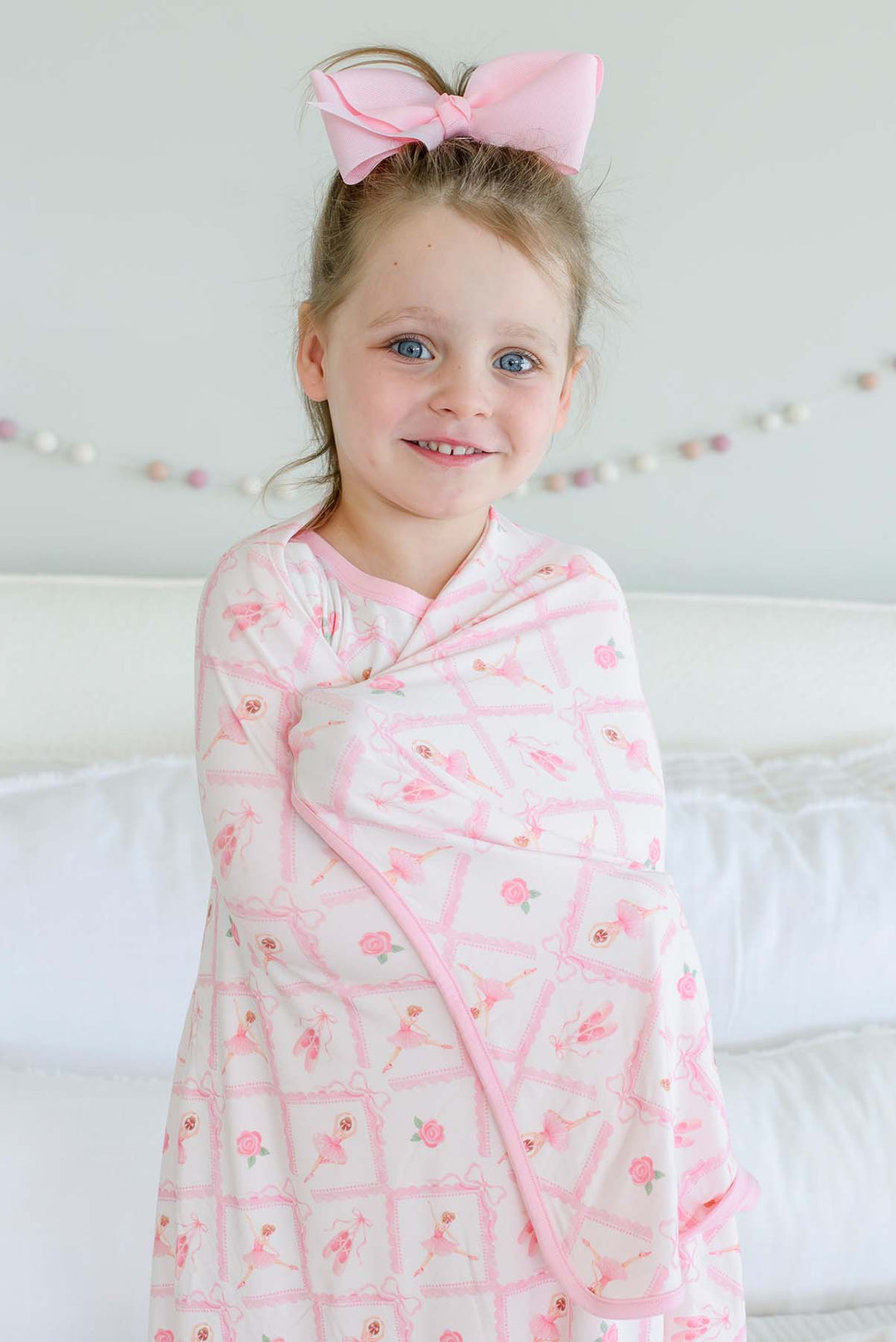 Smiling young girl standing on a bed with a pink bow in her hair, wrapped in a Caden Lane ballerina and roses pink print bamboo blanket