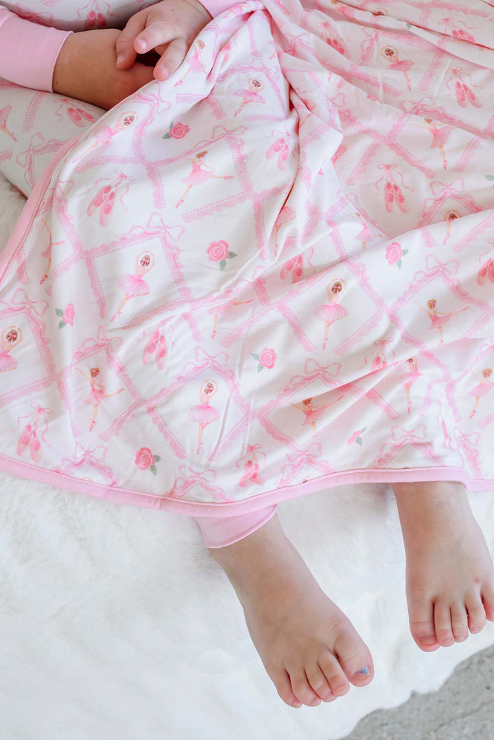 Smiling young girl sitting on a bed wearing Caden Lane ballerina and roses pink print pajamas, draped with a matching bamboo blanket