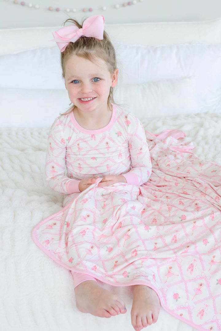 Smiling young girl sitting on a bed wearing Caden Lane ballerina and roses pink print pajamas, draped with a matching bamboo blanket