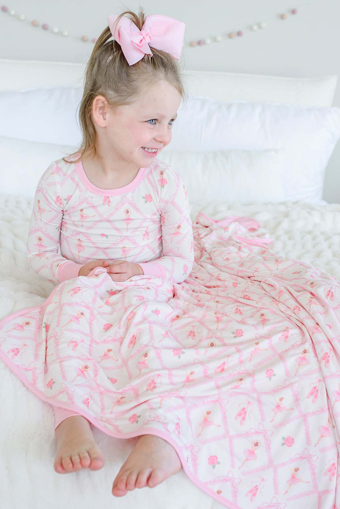 Smiling young girl sitting on a bed wearing Caden Lane ballerina and roses pink print pajamas, draped with a matching bamboo blanket