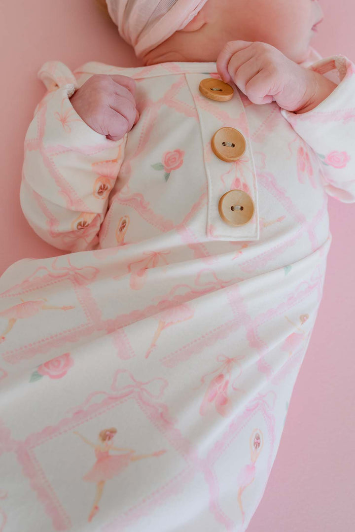 Newborn baby lying on a pink backdrop wearing a Caden Lane ballerina and roses pink print knotted gown with wooden buttons and matching oversized bow headband