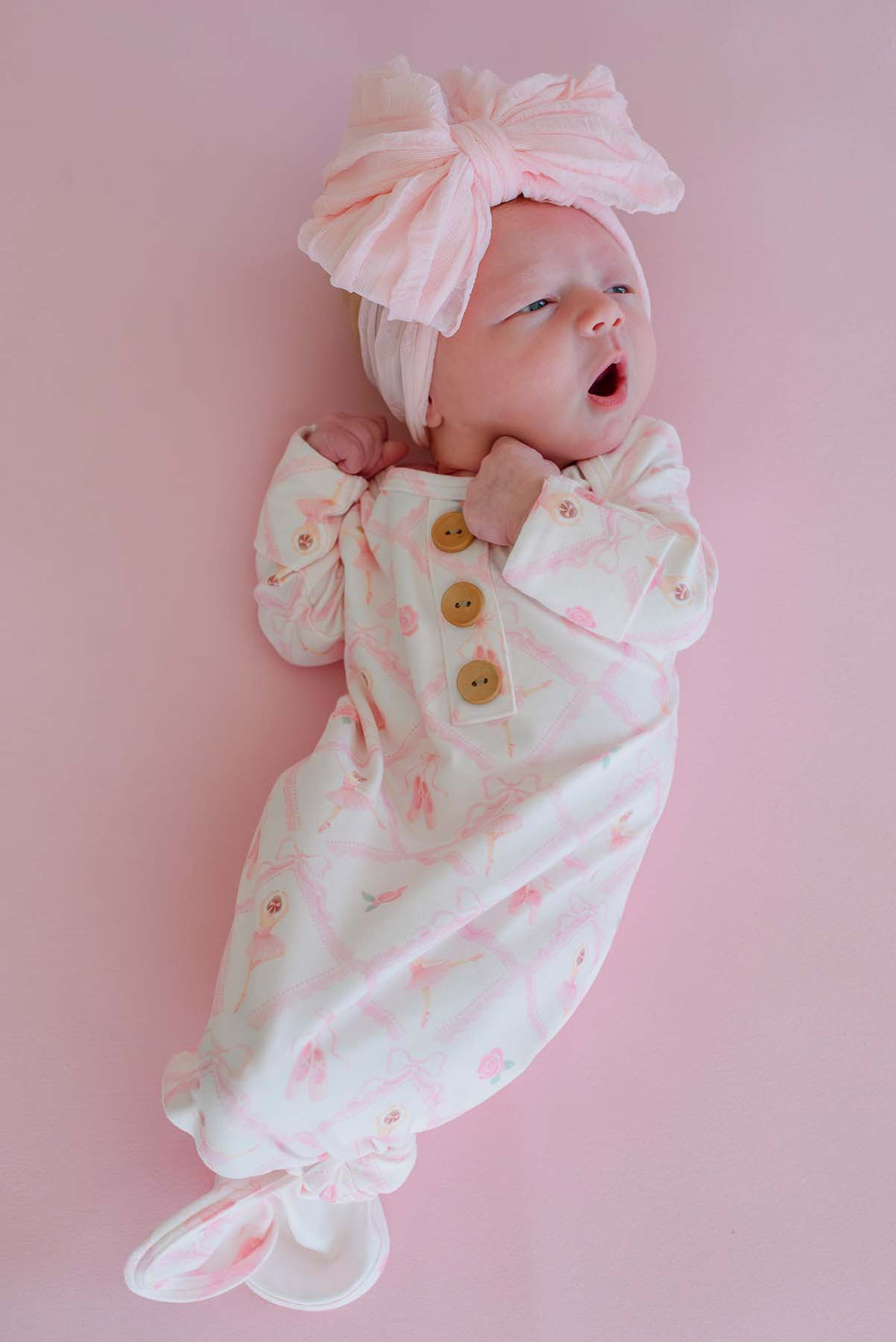 Newborn baby lying on a pink backdrop wearing a Caden Lane ballerina and roses pink print knotted gown with wooden buttons and matching oversized bow headband