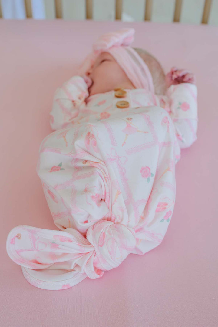 Newborn baby lying on a pink backdrop wearing a Caden Lane ballerina and roses pink print knotted gown with wooden buttons and matching oversized bow headband