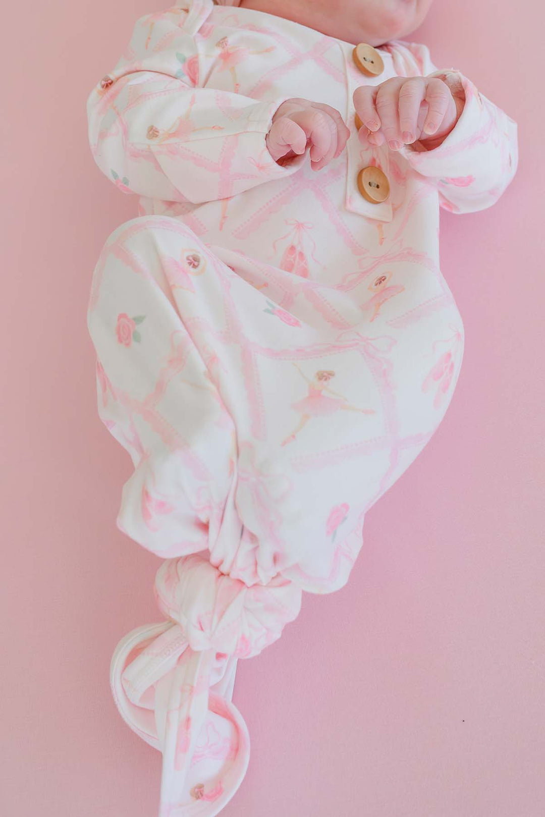 Newborn baby lying on a pink backdrop wearing a Caden Lane ballerina and roses pink print knotted gown with wooden buttons
