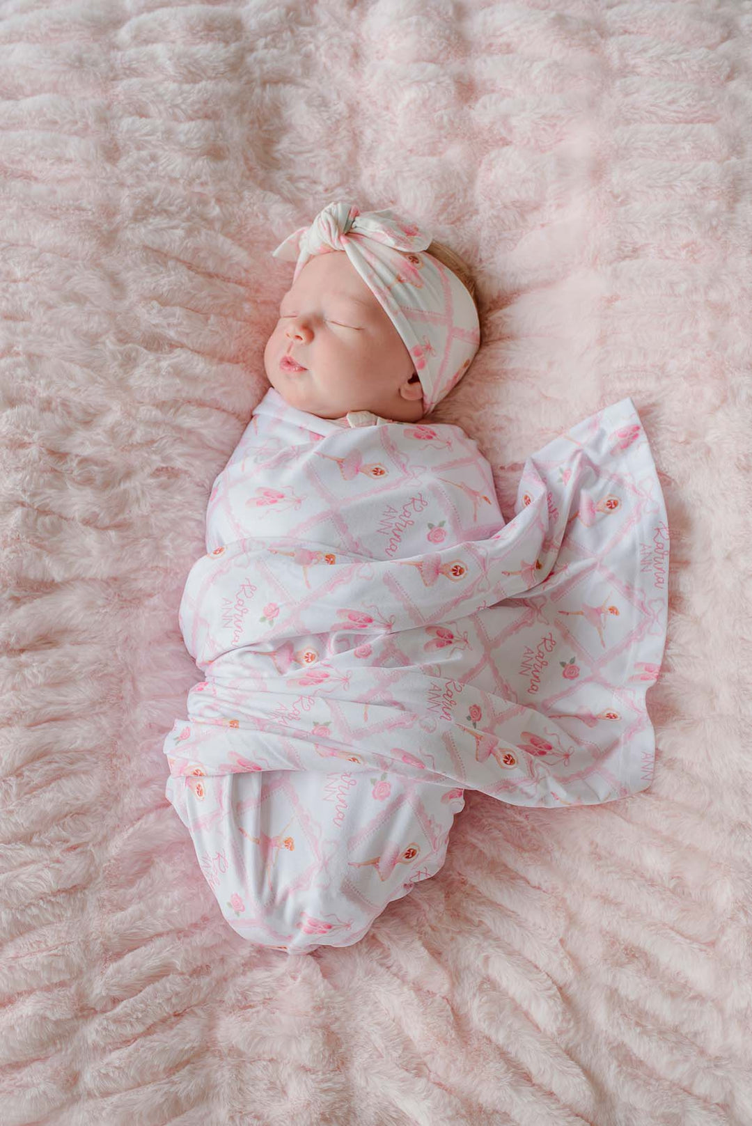 Close-up of sleeping newborn baby wearing a Caden Lane ballerina and roses pink print swaddle blanket with matching bow headwrap, laying on a pink faux fur blanket