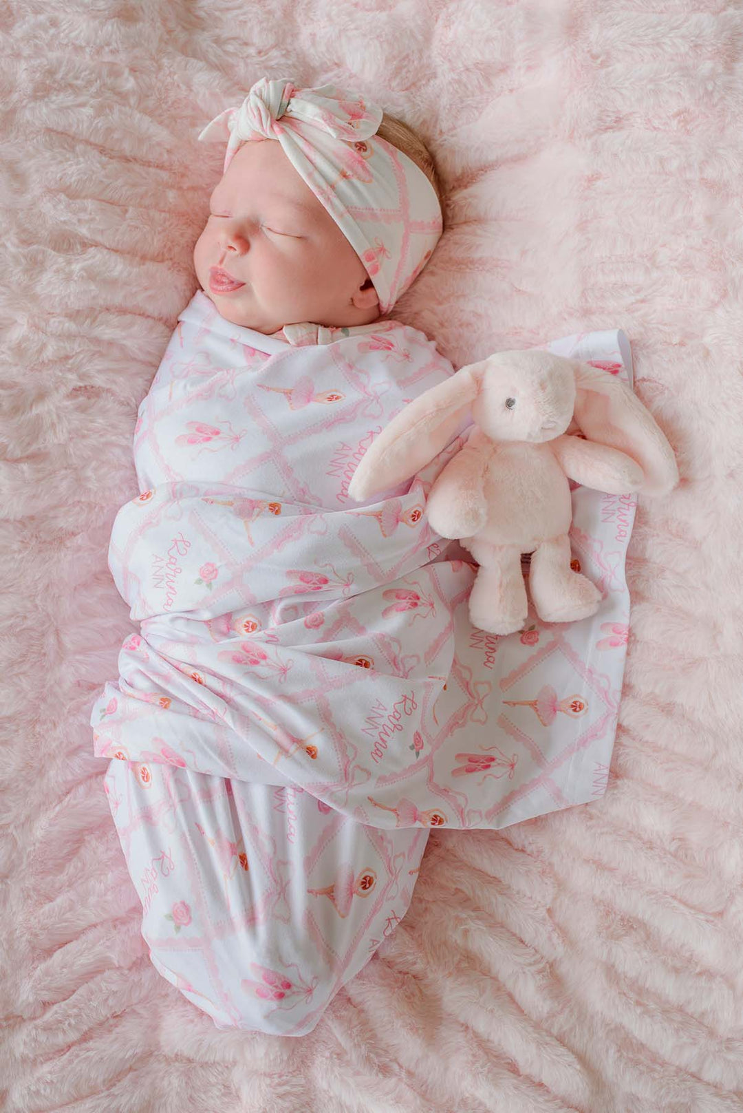 Sleeping newborn swaddled in a Caden Lane ballerina and roses pink print personalized swaddle blanket with matching knotted headband, on a pink fur blanket