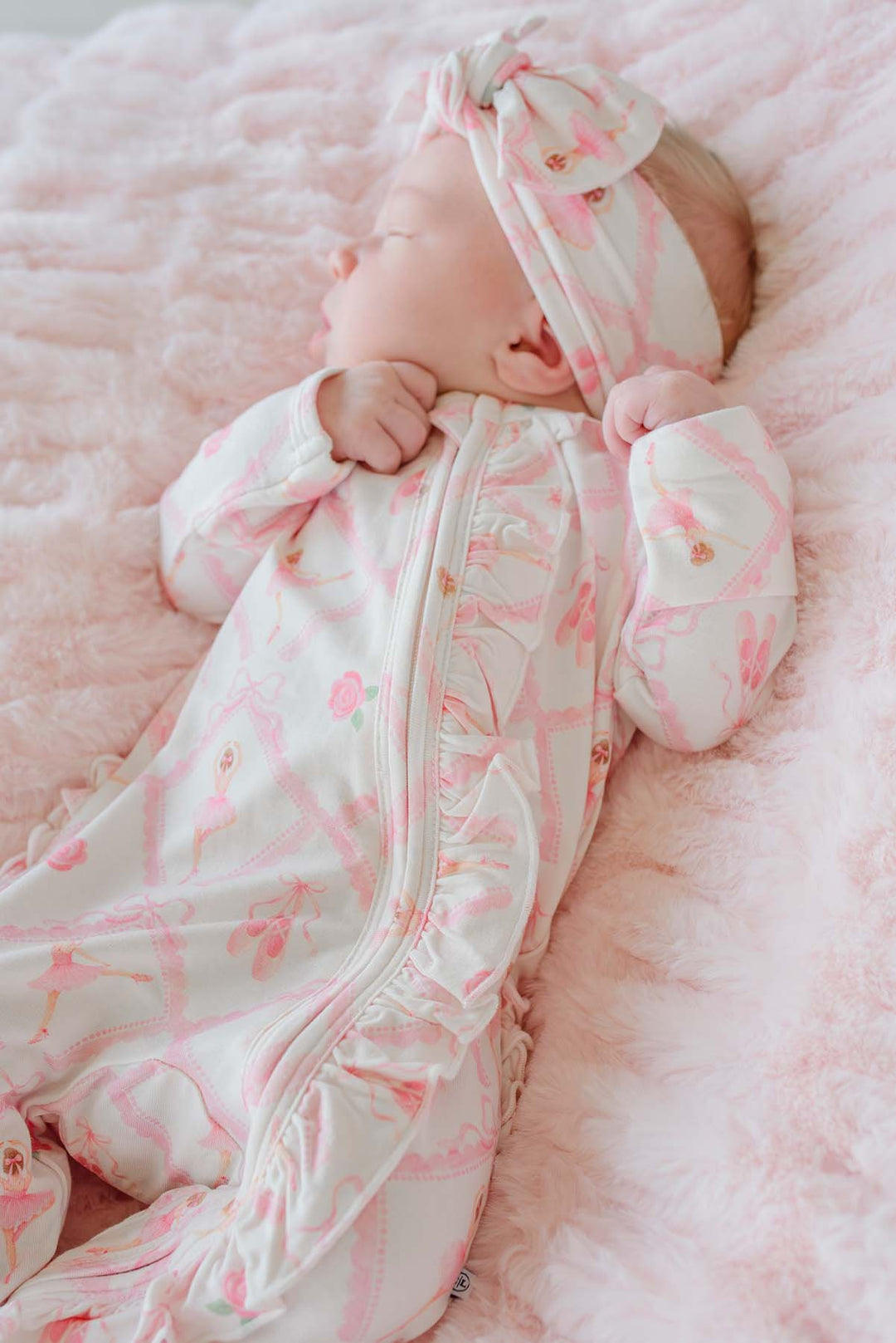 Sleeping newborn baby girl wearing a Caden Lane ballerina and roses pink print ruffle footie pajama with matching knotted headband, laying on a pink faux fur blanket