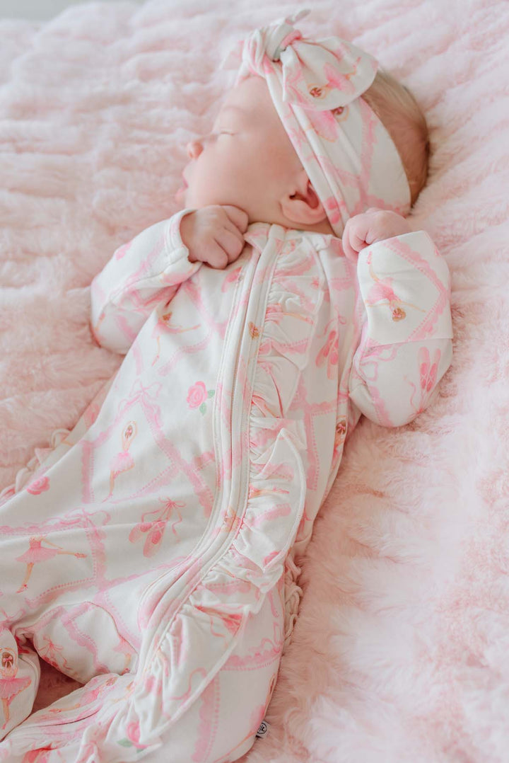 Sleeping newborn baby girl wearing a Caden Lane ballerina and roses pink print ruffle footie pajama with matching knotted headband, laying on a pink faux fur blanket