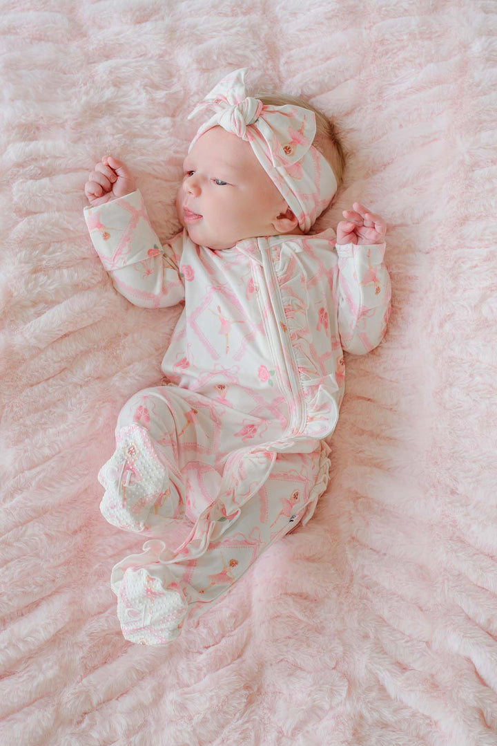 Sleeping newborn baby girl wearing a Caden Lane ballerina and roses pink print ruffle footie pajama with matching knotted headband, laying on a pink faux fur blanket