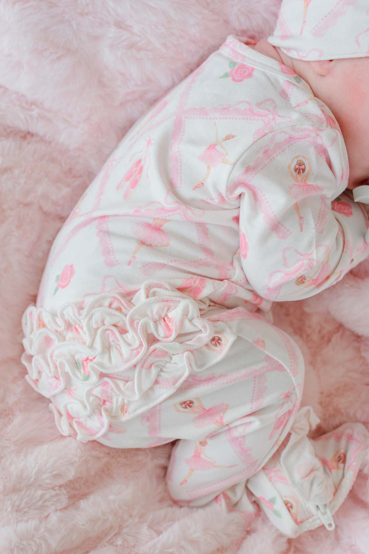 Sleeping newborn baby girl wearing a Caden Lane ballerina and roses pink print ruffle footie pajama with matching knotted headband, laying on a pink faux fur blanket