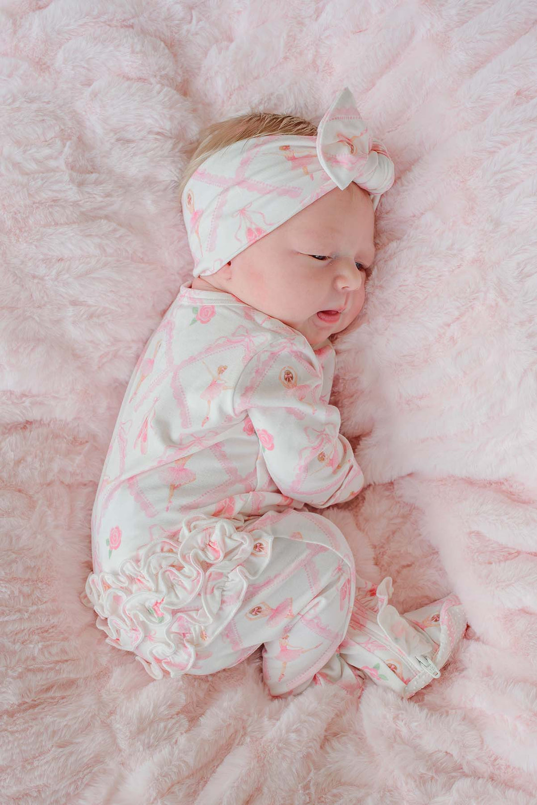 Newborn baby girl wearing a Caden Lane ballerina and roses pink print ruffle footie pajama with matching knotted headband, laying on a pink faux fur blanket