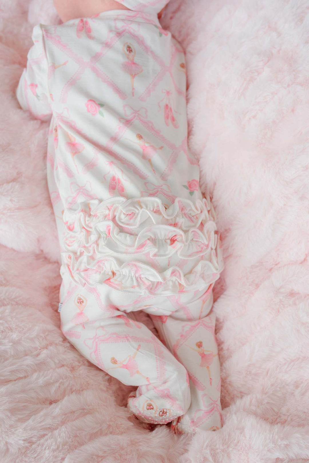 Newborn baby girl wearing a Caden Lane ballerina and roses pink print ruffle footie pajama with matching knotted headband, laying on a pink faux fur blanket