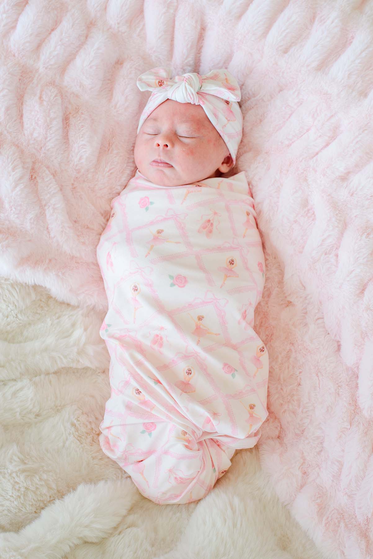 Sleeping newborn baby swaddled in a Caden Lane ballerina and roses pink print swaddle blanket with matching bow headwrap, laying next to a stuffed bunny on a pink fur blanket